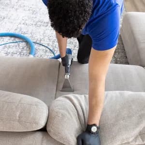 Worker wearing gloves cleans pale sofa upholstery with a vacuum hose attachment