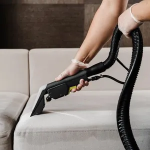 Person holding upholstery cleaning nozzle against pale sofa cushion beside dark backdrop