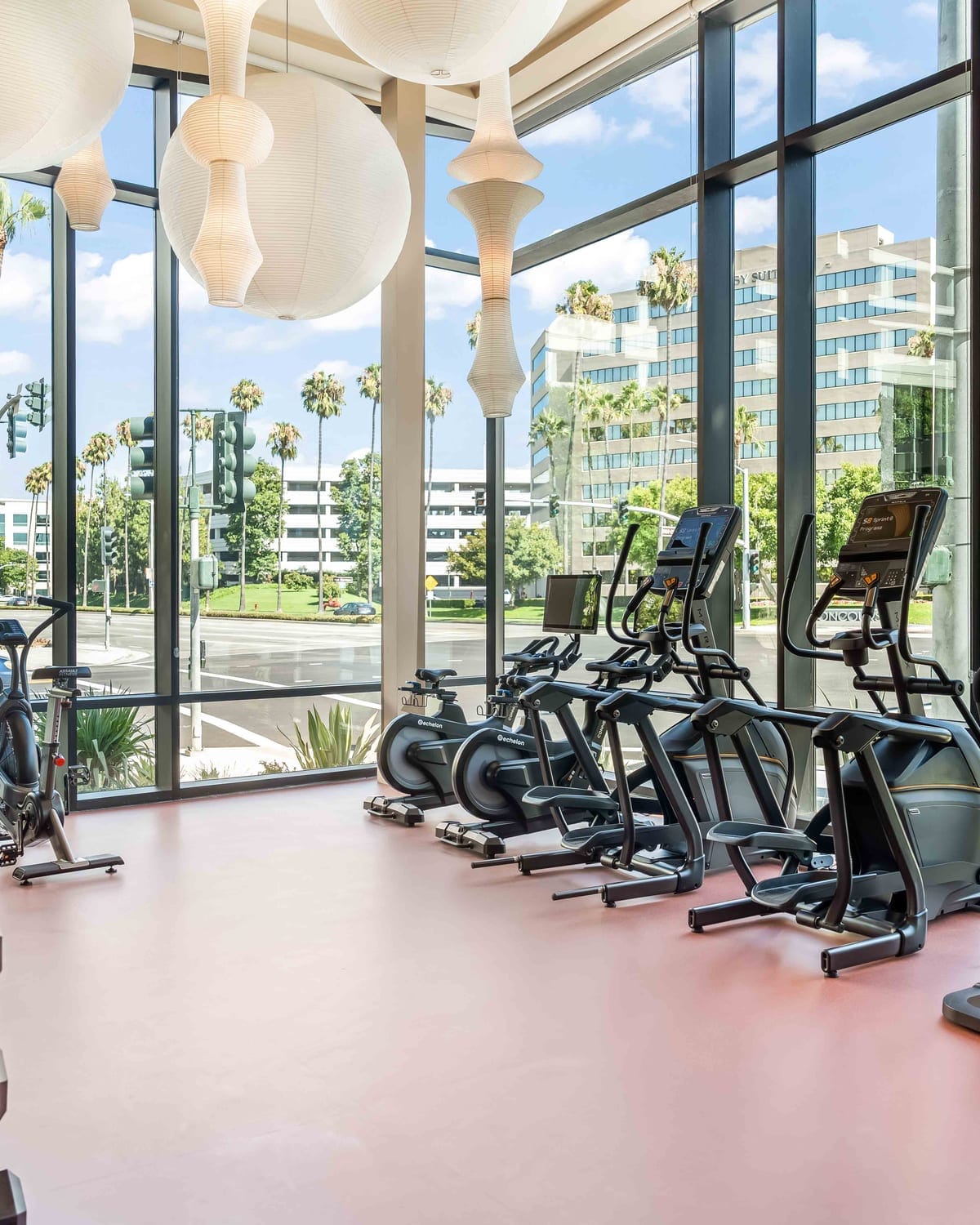 A building fitness center with cardio equipment along a full-height glass wall looking onto palm trees