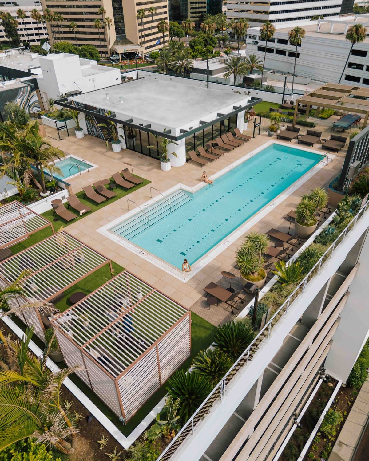 A rooftop pool deck seen from above, with a lap pool, cabanas and loungers