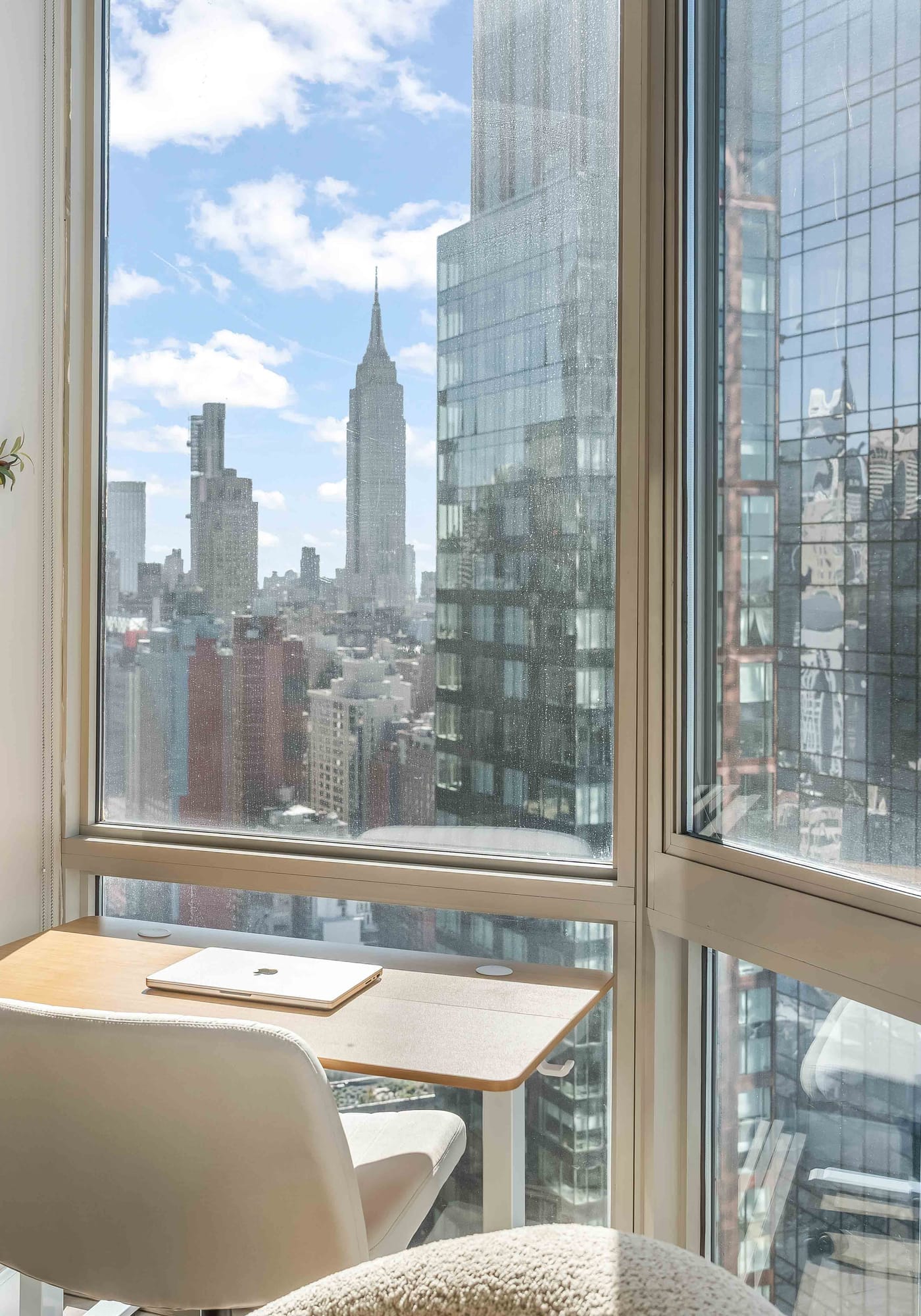 A desk and chair at the corner window of a World Class residence, a closed laptop on the desk and the Manhattan skyline beyond