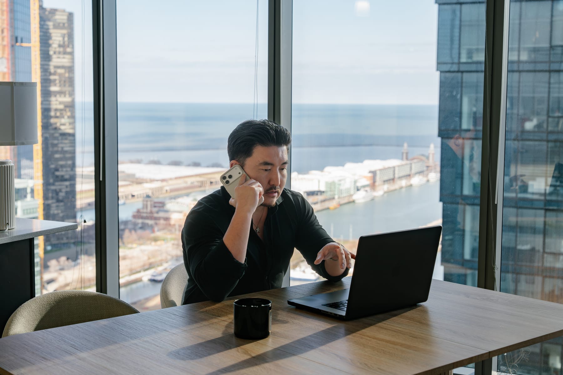A resident taking a call and working on a laptop at a dining table, with a waterfront city view behind