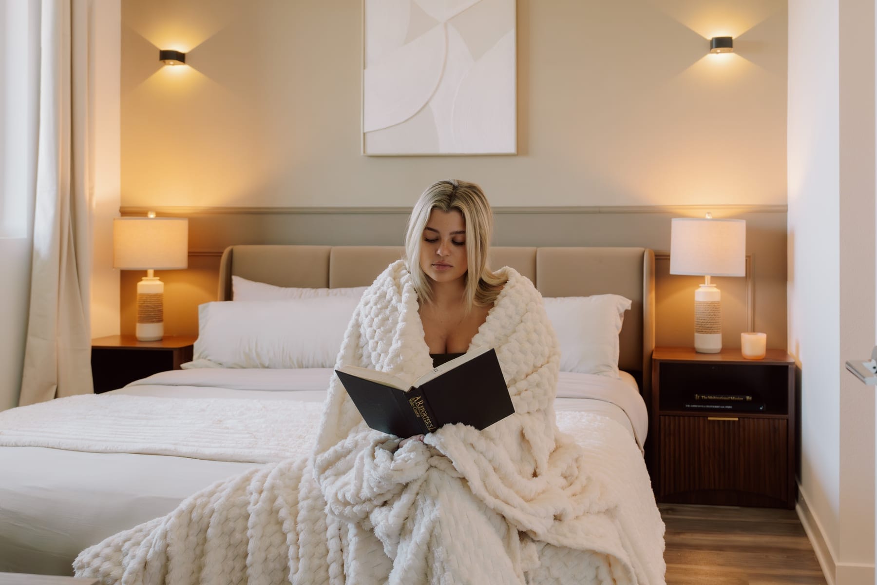 A resident resting in bed with a book, wrapped in a throw with the bedside lamps lit and the curtains drawn