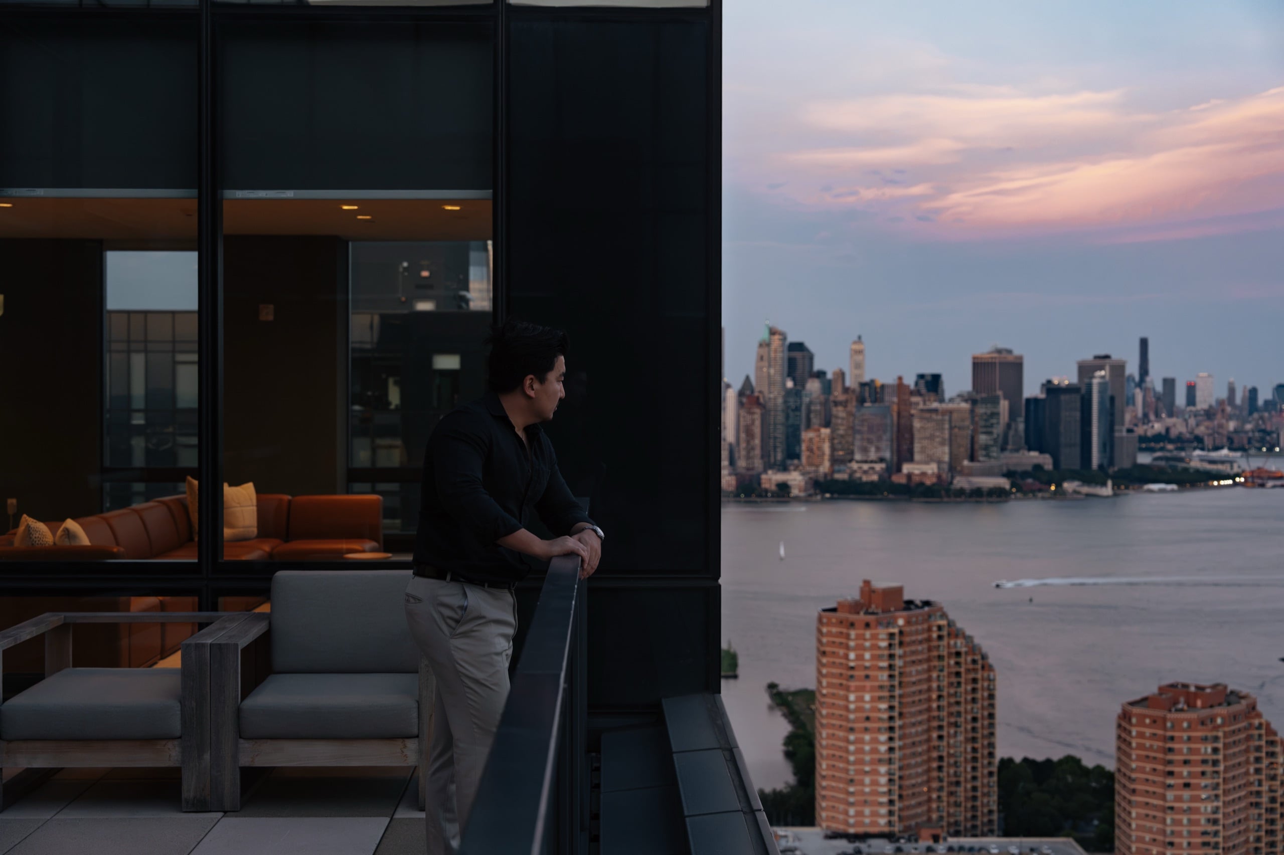 A resident leaning on the railing of a World Class terrace at dusk, looking across the Hudson to the lower Manhattan skyline