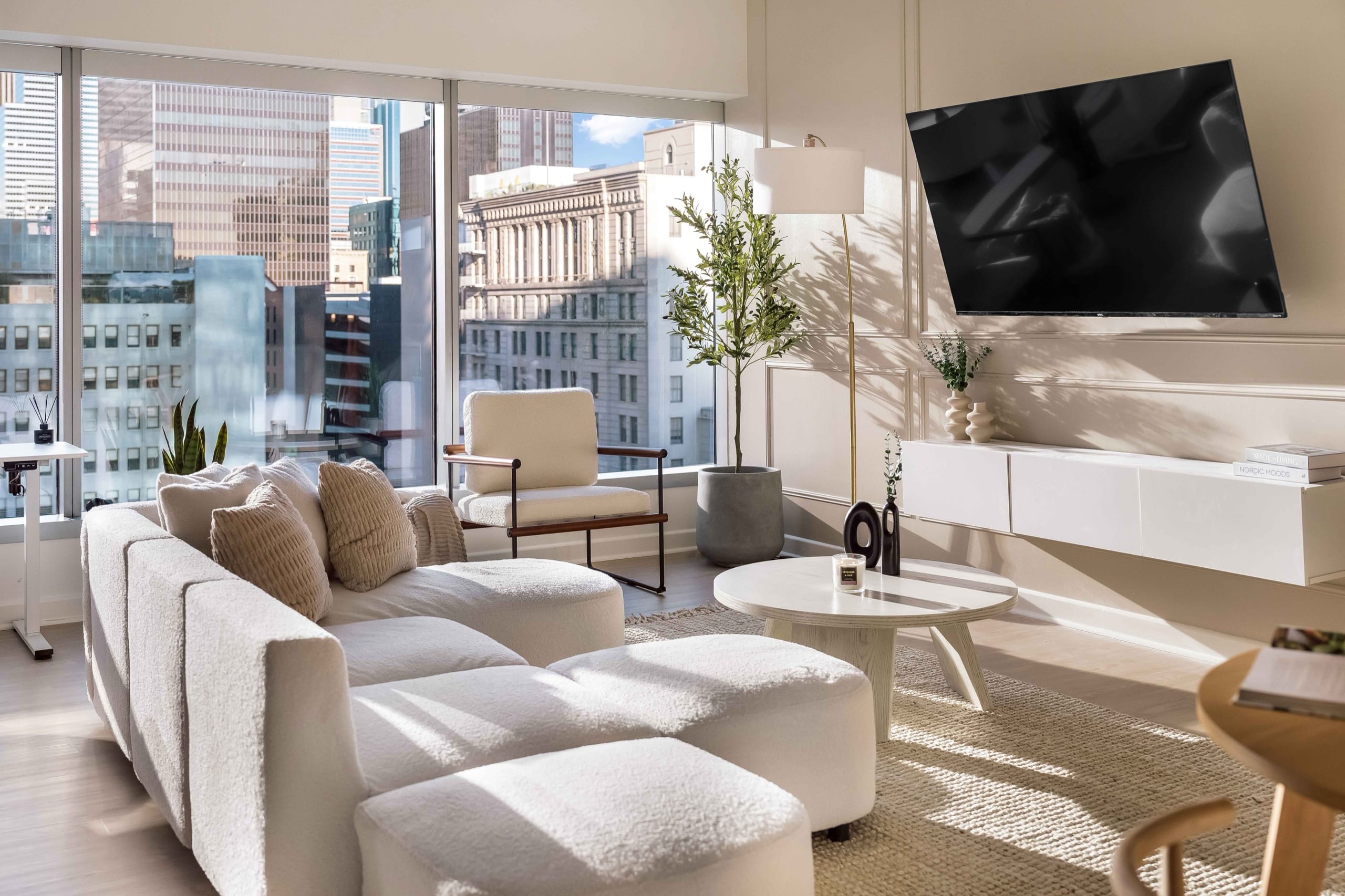 A World Class living room in afternoon light: a boucle sectional, an olive tree, a floating console and sculptural ceramics, with the city through full-height windows