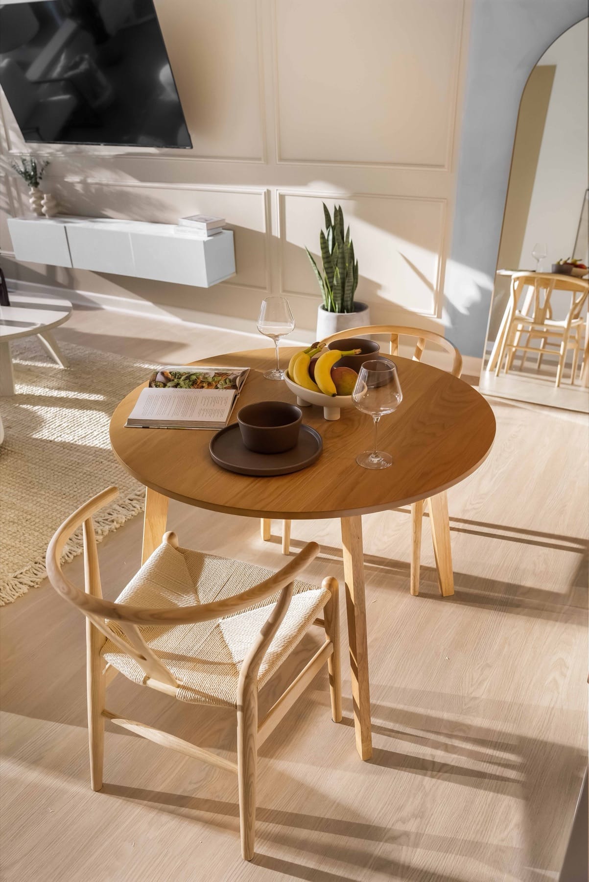 A sunlit dining nook in a World Class residence: an oak table with woven-seat chairs, an open cookbook, a bowl of fruit and glassware