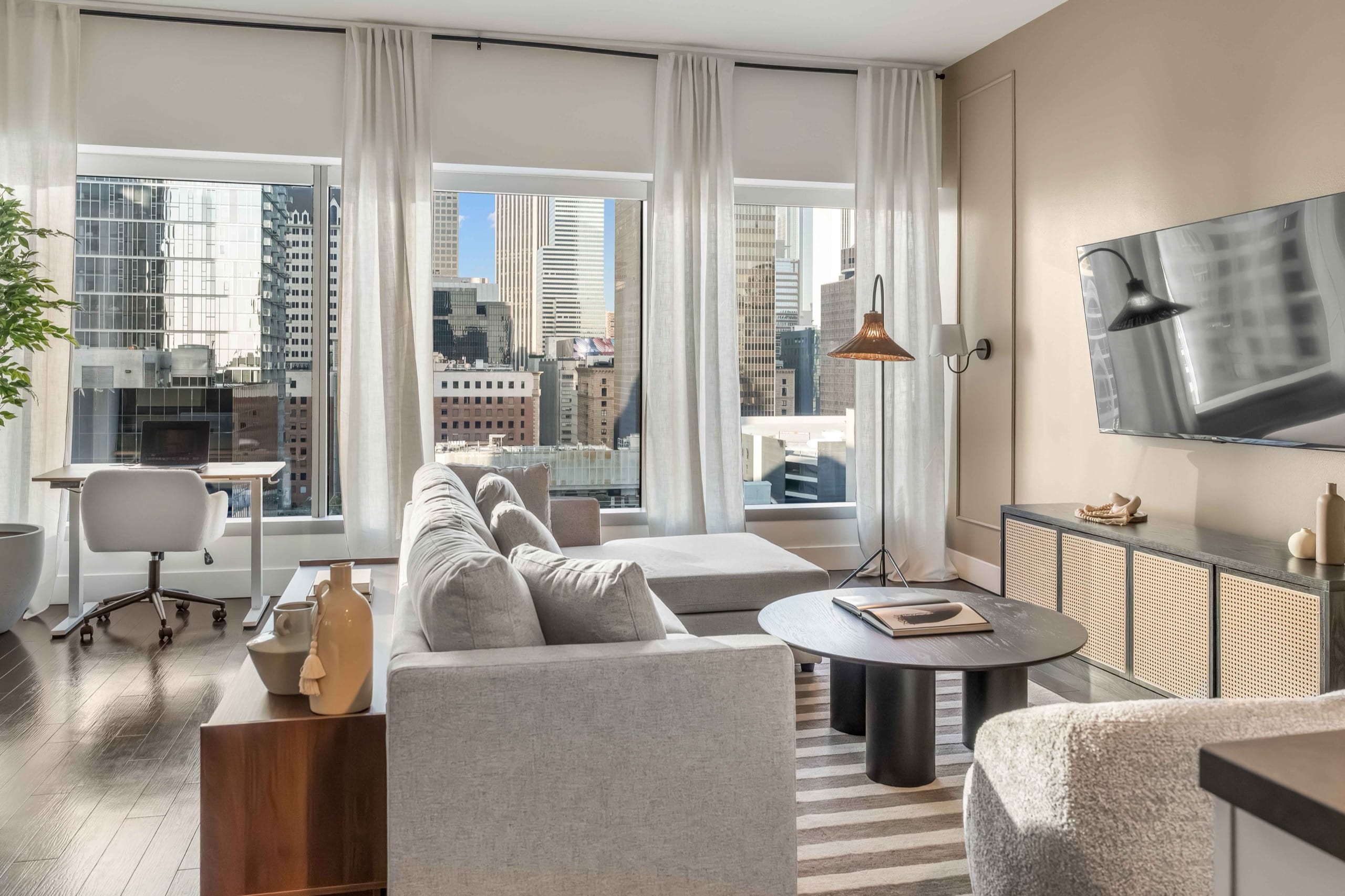 The living room of a World Class residence, with sheer curtains across full-height windows, a pale sectional, a cane sideboard and the Midtown skyline beyond