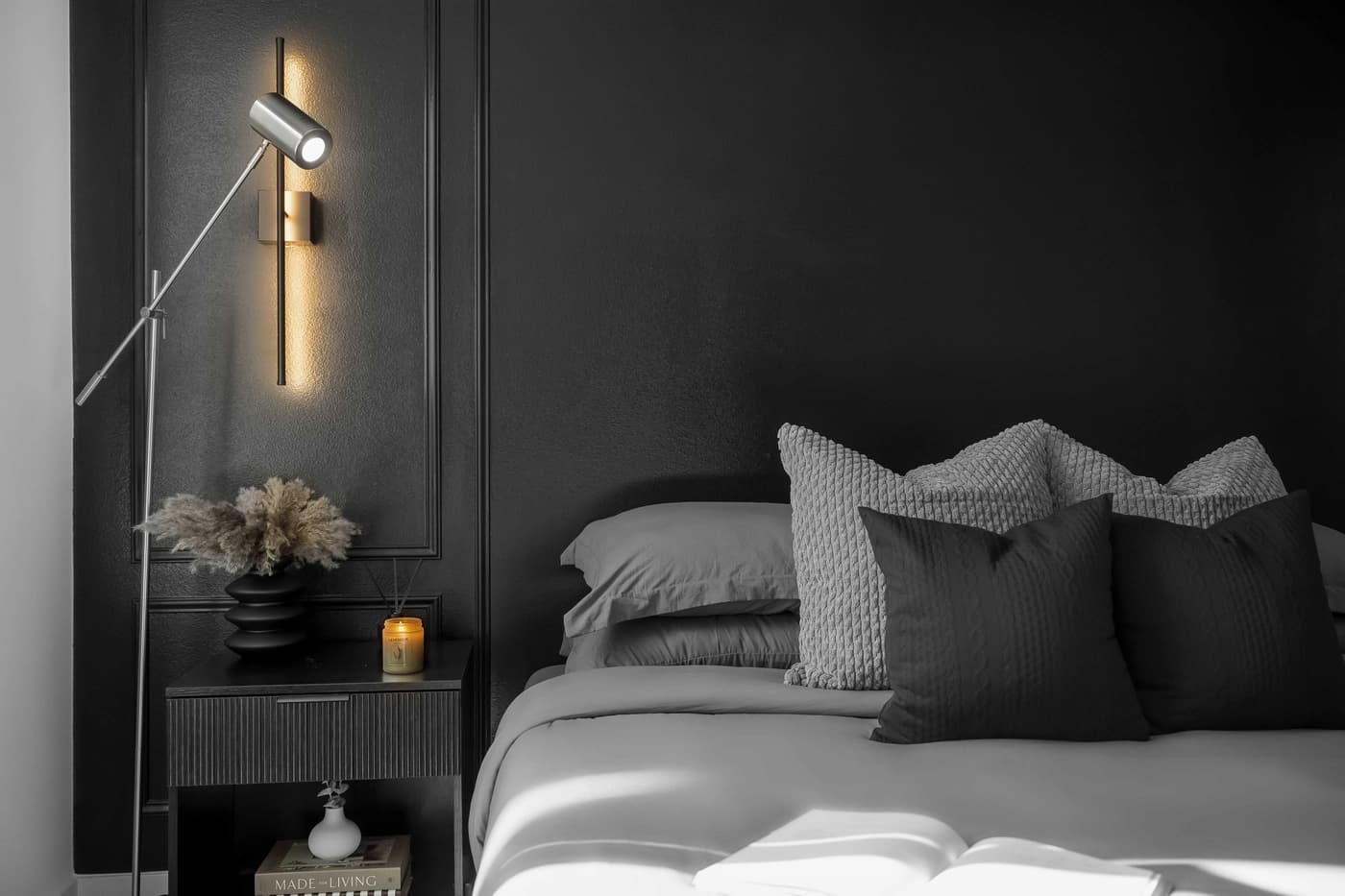 Bed with dark pillows beside black nightstand and wall-mounted reading light against dark paneled walls at The Alexander Condo in Santa Ana