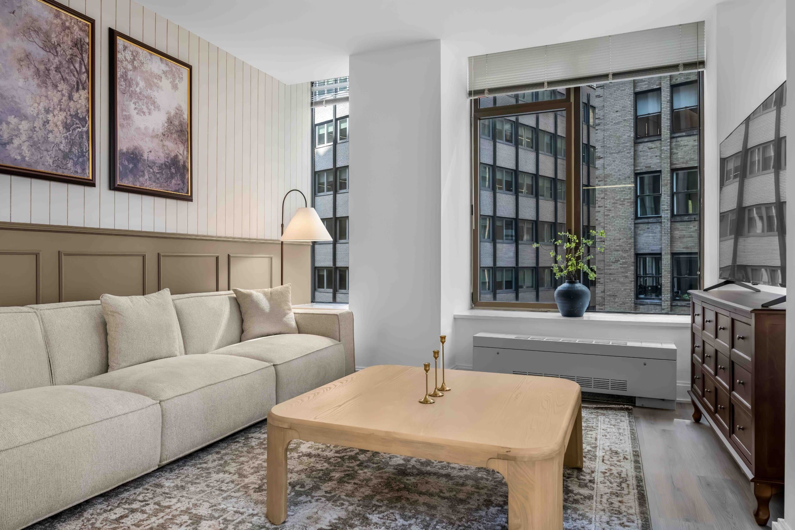 Living room pale sofa and wooden table beside television and tall building-view windows at Casa Loma by World Class® in New York