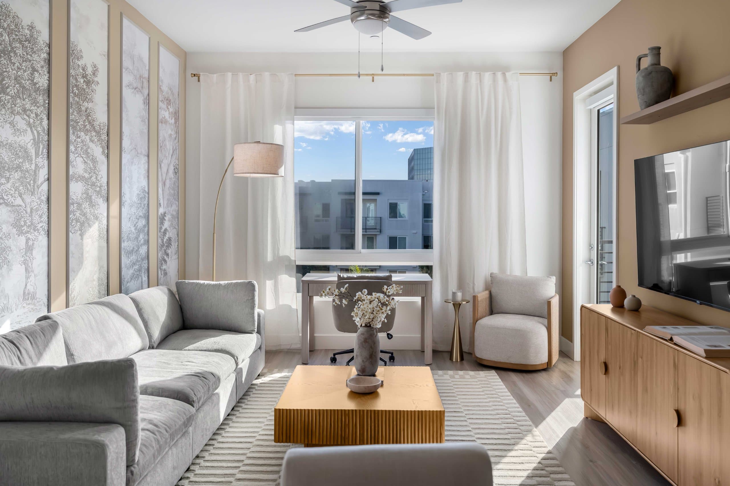 Living room with gray sofa, wooden table, television, cabinet, chairs, plants, and sheer-curtained window at The Seraphina in Newport Beach