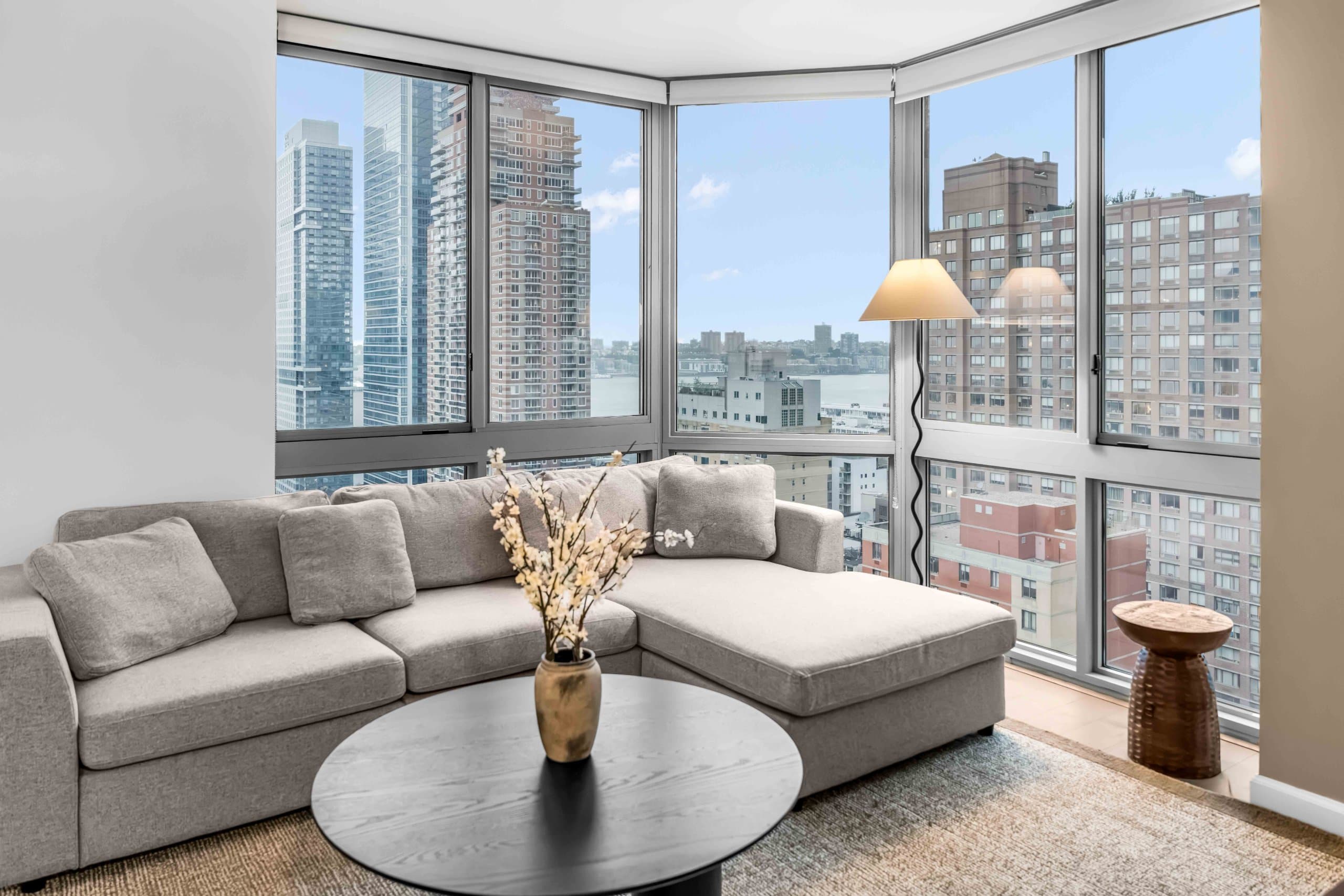 Gray sectional sofa and round black table with flowering vase beside corner city-view windows at The Chelsea Highrise by World Class® in New York City